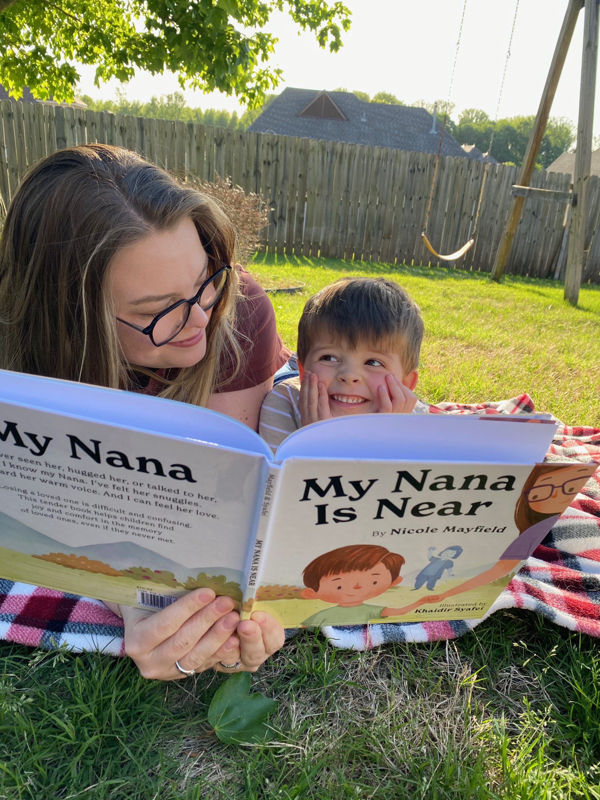 Author Nicole Mayfield reads My Nana Is Near with son, Noah, while lying on a blanket in the grass.