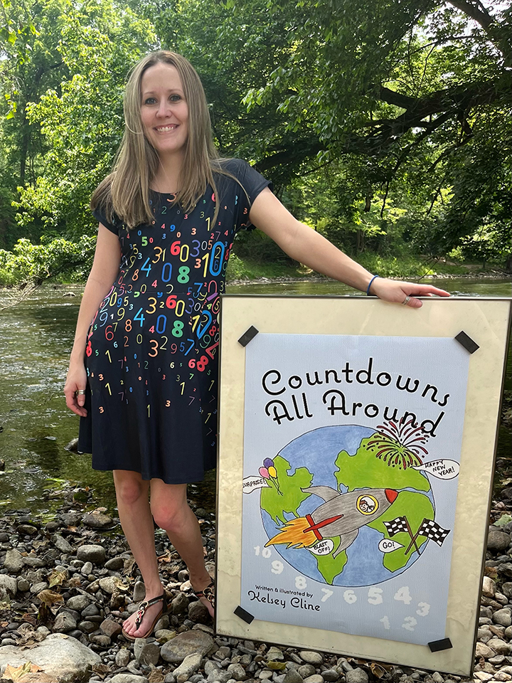 Kelsey Cline, author of Countdowns All Around, stands with a poster of her book cover beside a creek.